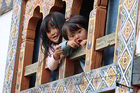 Two curious children peek from a wooden window, capturing the gentle charm of daily life in Paro, Bhutan