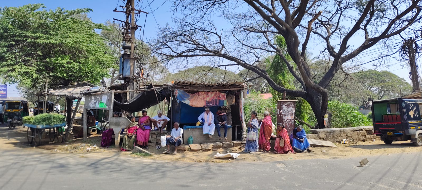 ತಂಗುದಾಣವಿಲ್ಲದೆ ಮರದ ಕೆಳಗೆ ಕುಳಿತು ಬಸ್‌ಗಾಗಿ ಕಾಯುತ್ತಿದ್ದ ಪ್ರಯಾಣಿಕರು