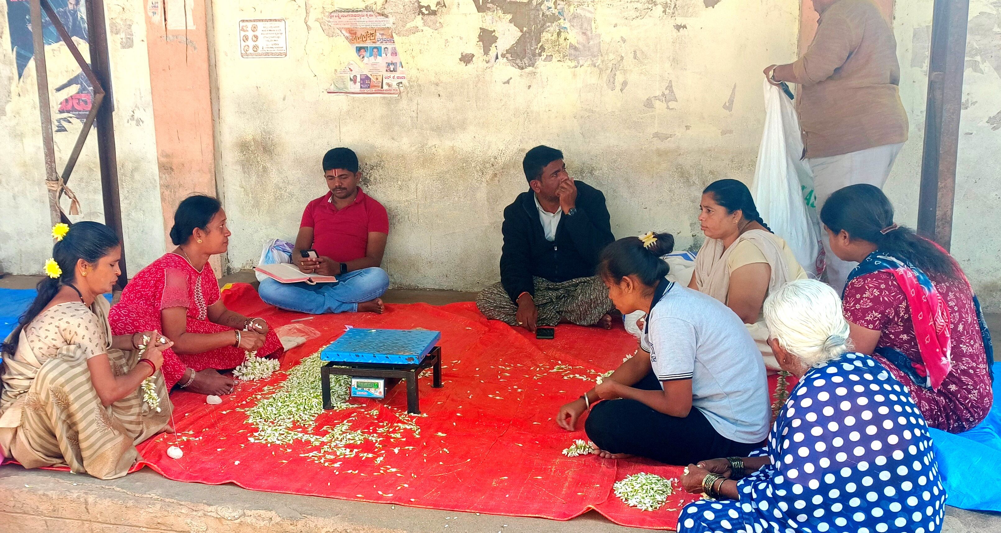 ಮಹಿಳೆಯರು ವ್ಯಾಪಾರಿಯಿಂದ ಕೊಂಡು ಹೂವು ಕಟ್ಟುತ್ತಿರುವುದು