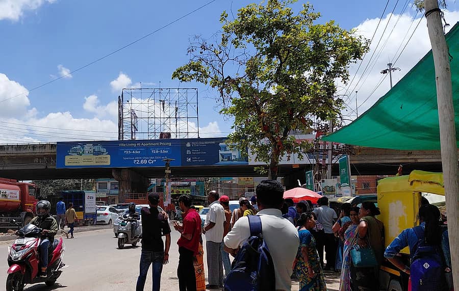 ಬೂದಿಗೆರೆ ಕ್ರಾಸ್ ಬಳಿ ಬಸ್ ತಂಗುದಾಣವಿಲ್ಲದೆ ಪ್ರಯಾಣಿಕರು ಮರವನ್ನು ಆಶ್ರಯಿಸಿರುವುದು