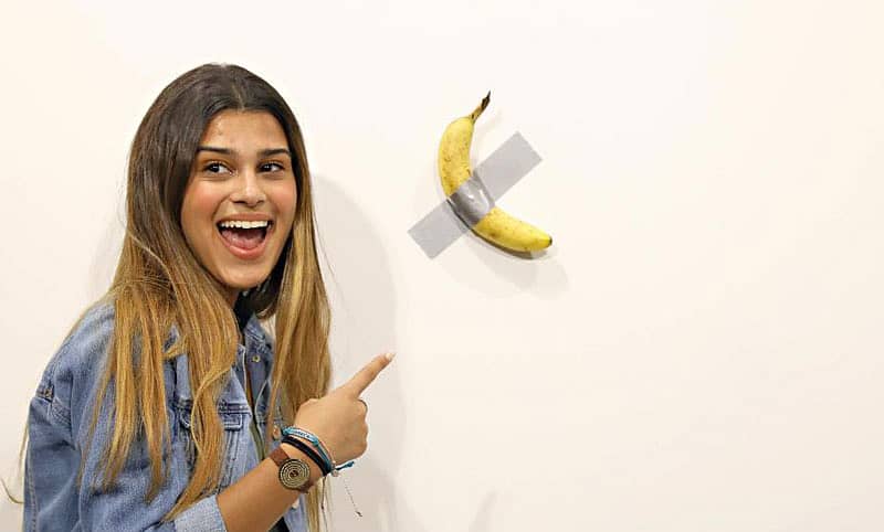 ಬಾಳೆಹಣ್ಣಿನ ಕಮೀಡಿಯನ್ ಕಲಾಕೃತಿಯ ಎದುರು ನಿಂತು ಫೋಟೊ ಪಡೆಯುತ್ತಿರುವ ಯುವತಿ