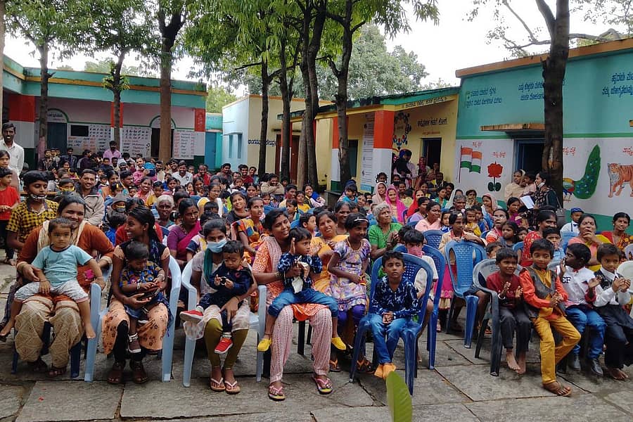 ಸುಗಟೂರು ಗ್ರಾಮದ ಶಾಲಾವರಣದಲ್ಲಿ ಹೊಸ ಉಡುಪು ಧರಿಸಿ ಪೋಷಕರೊಂದಿಗೆ ಕುಳಿತಿರುವ ಮಕ್ಕಳು