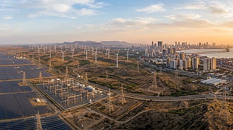 An aerial view of solar farms, wind turbines, and electricity transmission infrastructure near a modern city skyline.