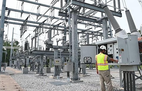 Avangrid field worker inspecting control equipment at an electrical substation, with transformers, circuit breakers and overhead grid infrastructure visible.