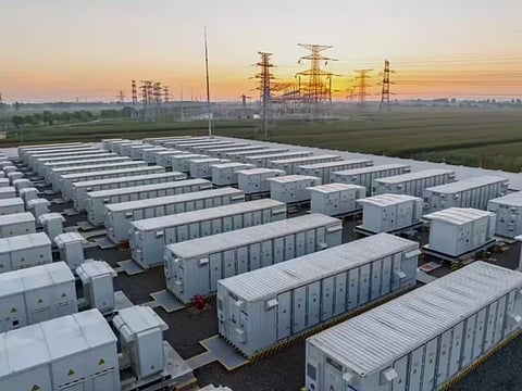 Rows of containerized battery energy storage units at a large-scale facility, with electrical substations and high-voltage transmission towers visible in the background.