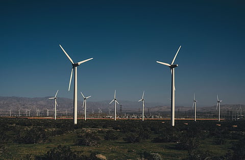 Multiple wind turbines operating across an expansive onshore wind farm, with mountains and power infrastructure visible in the background.