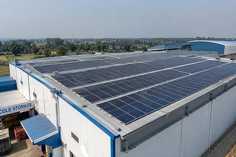 Large rooftop solar panel arrays installed on a cold storage facility, with agricultural fields, trees and rural buildings visible in the surrounding West Bengal landscape.