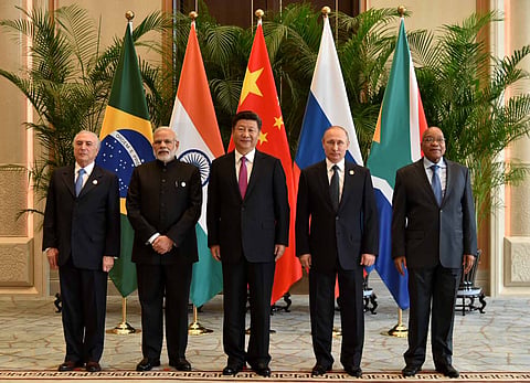 Five BRICS leaders standing together in front of the national flags of Brazil, India, China, Russia and South Africa at a formal summit venue.