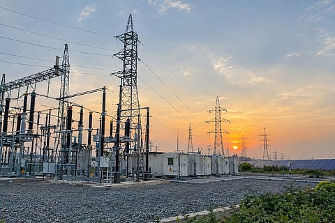 Electrical substation with transformers, transmission towers and overhead power lines, with a solar installation visible in the background.