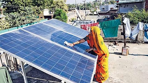 A woman wearing a yellow and orange sari cleans blue rooftop solar panels on a residential terrace in Modhera village, Gujarat, with nearby houses, trees and laundry visible in the background.
