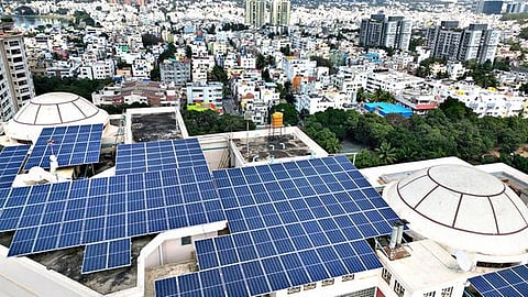 Blue solar panels cover the rooftop of a building amid a densely populated urban landscape in Karnataka, India.