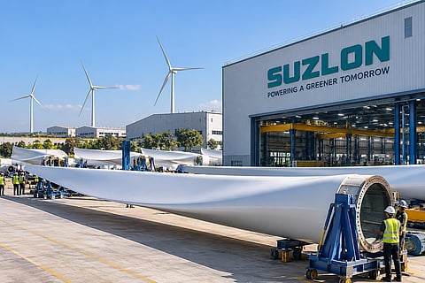 Large wind turbine blades outside a modern Suzlon-branded manufacturing facility, with industrial equipment, workers and wind turbines in the background.