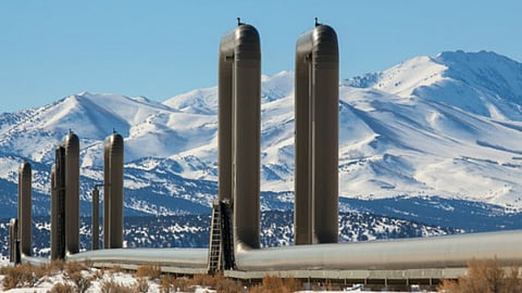 Tall cylindrical geothermal facility structures and connected pipelines in a snow-covered landscape, with snow-capped mountains in the background.