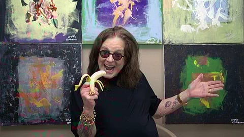 Ozzy Osbourne smiling with a banana in front of abstract paintings