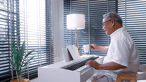 Older man seated at a piano holding a cup of tea in a bright, modern home