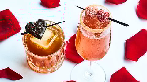 Two Valentine’s cocktails with heart garnishes and rose petals on a white tablecloth