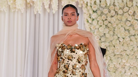 Model walks in a strapless gold gown with 3D floral appliqué and sheer tulle cape backdrop