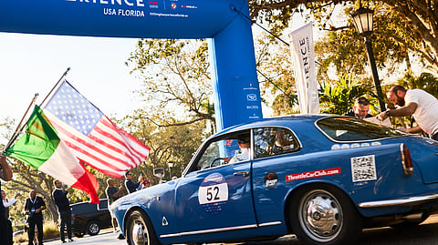Blue Alfa Romeo race car number 52 driving past start line with Italian and U.S. flags waving
