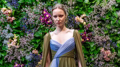 Model in flowing sage and blue chiffon evening gown before floral wall in Milan