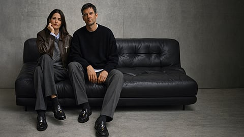Male and female models on a black leather sofa wear gray tailoring and black loafers