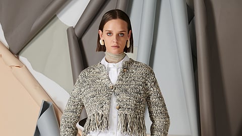 Model in fringed knit jacket and wide leg cream trousers against sculptural studio backdrop