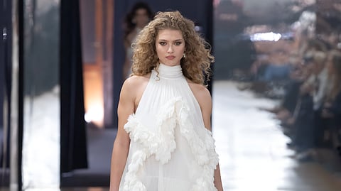 Model walks in a flowing ivory halter gown with layered ruffle trim and black thigh-high boots