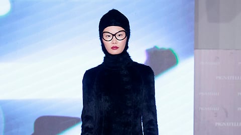 Model in black textured dress with headpiece and glasses walking runway through fog