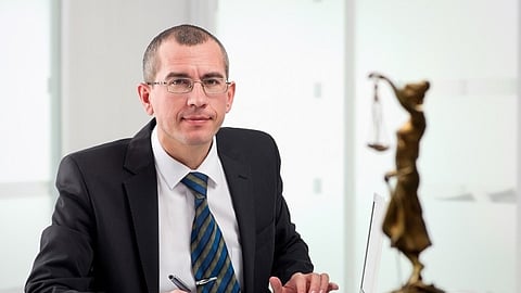 Man in suit sitting at desk with legal documents and scales of justice in foreground