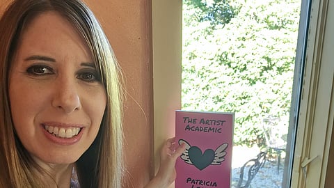 Patricia Leavy holding The Artist Academic beside a window with writing materials