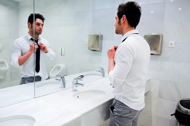 A man adjusting his tie in front of a mirror within a public restroom. 