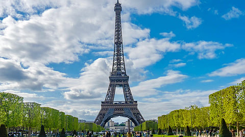The Eiffel Tower rises above Paris under a bright sky with visitors on the Champ de Mars lawn