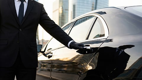 A chauffeur in a black suit and gloves opens the rear door of a black luxury sedan at the curb