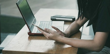 Person's hands using a smartphone next to an open laptop on a wooden desk.