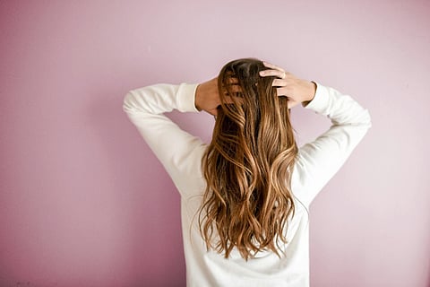 Woman with long hair standing against a pink wall, holding her head with both hands