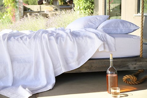 Outdoor bed with white linens, pillows, and a glass bottle beside a wooden platform