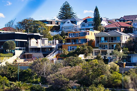 Residential neighborhood with modern houses, trees, and hillside landscape under a blue sky