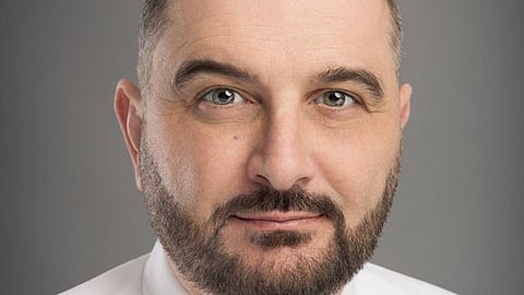 Professional headshot of a bearded man wearing a white shirt and purple tie.