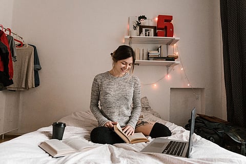 Woman reading on a bed beside an open laptop and books.