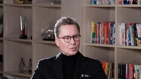 A man in a black suit is seated in a chair, looking confidently at the camera.
