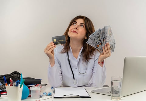 A woman in a white coat holds a credit card, smiling confidently at the camera.