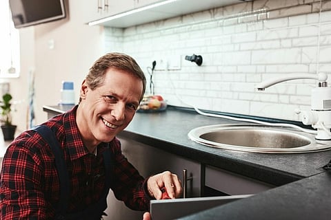 Smiling technician repairing a kitchen cabinet or sink area in a modern home.