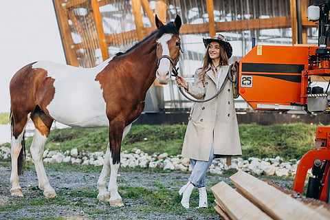 A smiling woman wearing a trench coat and cowboy hat holding the lead of a brown and white horse
