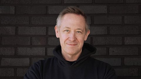 Smiling man in a "DOMINATE" hoodie against a dark brick wall.