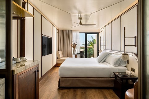 A hotel room with a king bed, wood floors, slatted wall panels and a terrace opening to palm trees