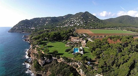 Aerial view of a low stone hotel on a green clifftop above the Mediterranean with mountains behind