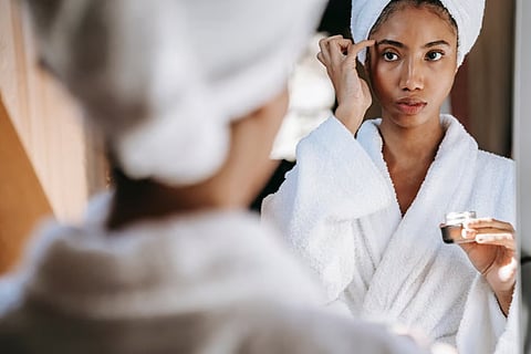 Woman in bathrobe applying skincare in mirror.