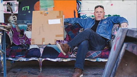 Man sitting on a patchwork couch next to a purple guitar.
