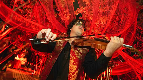 Male violinist in a gold and red brocade vest and masquerade mask plays under red lighting.