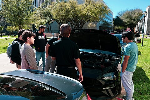 Group of people looking at a car engine outdoors.