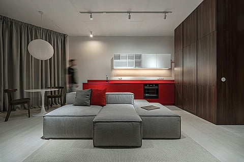 Modern living room with a gray sectional sofa and red kitchen counters.